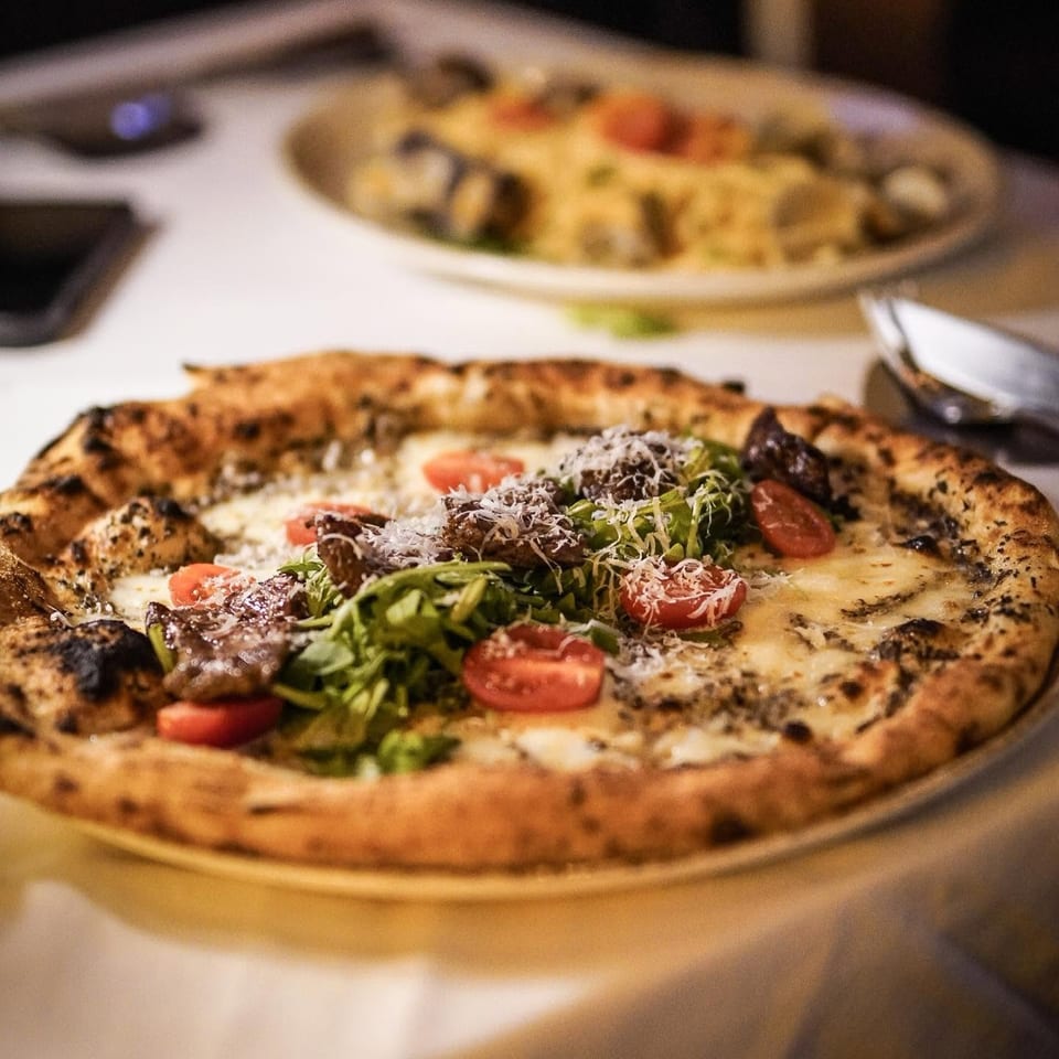 Wood-fired pizza topped with arugula, cherry tomatoes, roasted vegetables, and grated cheese, with another pizza in the background.