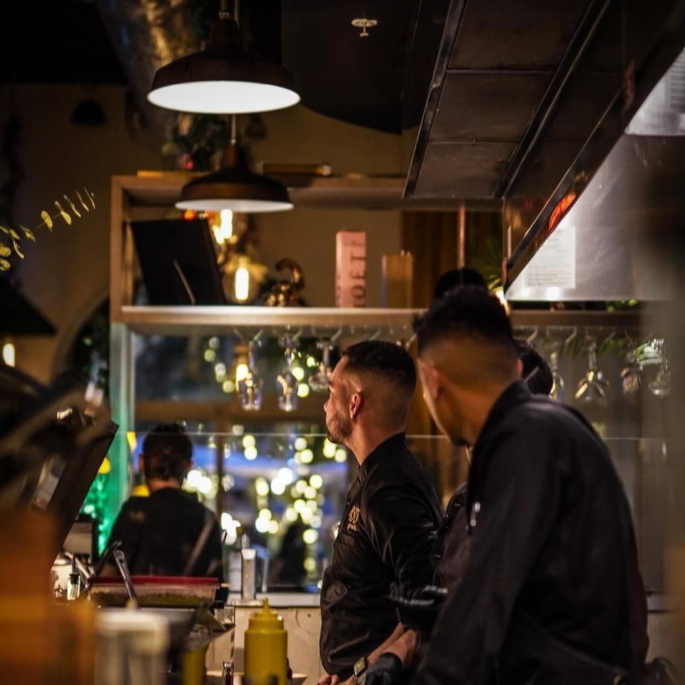 Restaurant staff work behind a counter beneath pendant lights, with kitchen equipment, condiments, and city lights visible through the window.