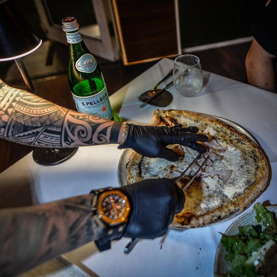 Gloved, tattooed servers carve a large baked fish at a restaurant table beside San Pellegrino water and side dishes.