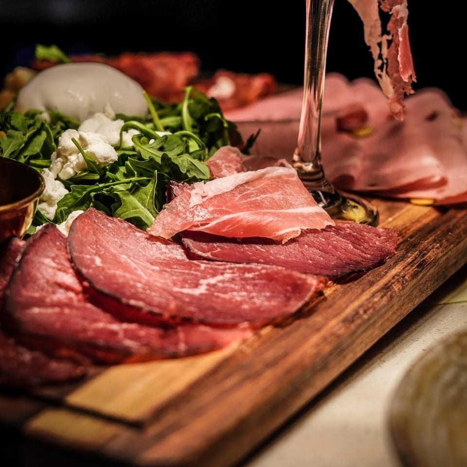 Wooden charcuterie board arranged with sliced cured meats, arugula, cauliflower, burrata, and vegetables.