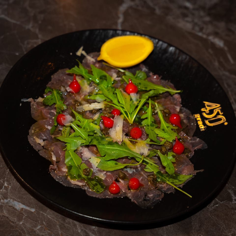 Thinly sliced beef carpaccio topped with arugula, red berries, capers, shaved cheese, olive oil, and lemon on a black plate.