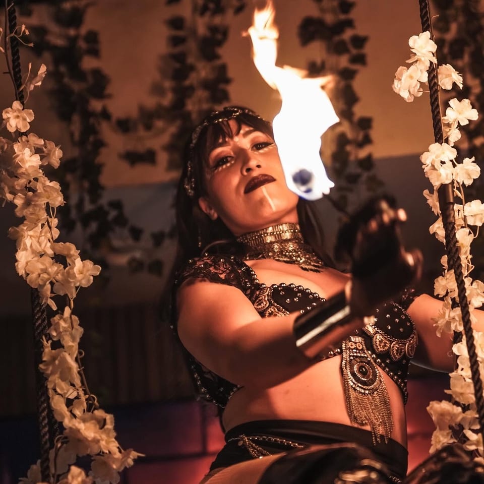 Entertainer in ornate black attire performs amid flames and flower-decorated ropes in a dim, theatrical setting.