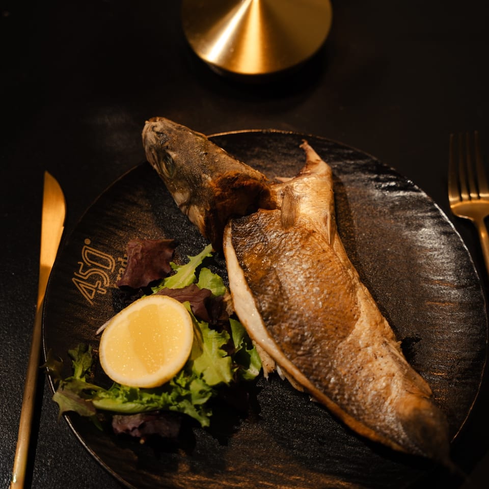 Whole roasted fish served on a black plate with leafy greens and lemon, alongside gold-toned cutlery in a dim restaurant setting.