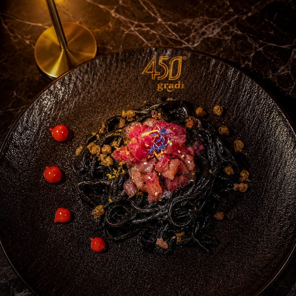 Elegant black pasta dish topped with colorful diced ingredients, tomatoes, and golden raisins on a 450 Gradi plate beside a brass lamp.