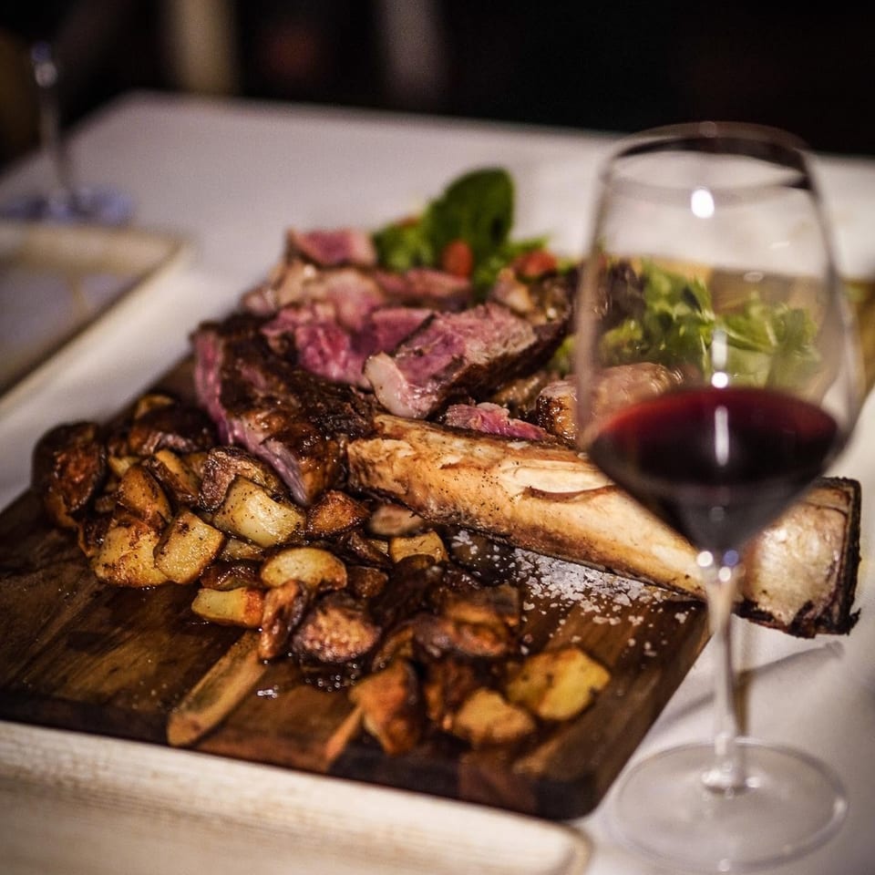 Sliced rare steak with roasted potatoes, vegetables, and red wine served on a wooden board at a restaurant table.