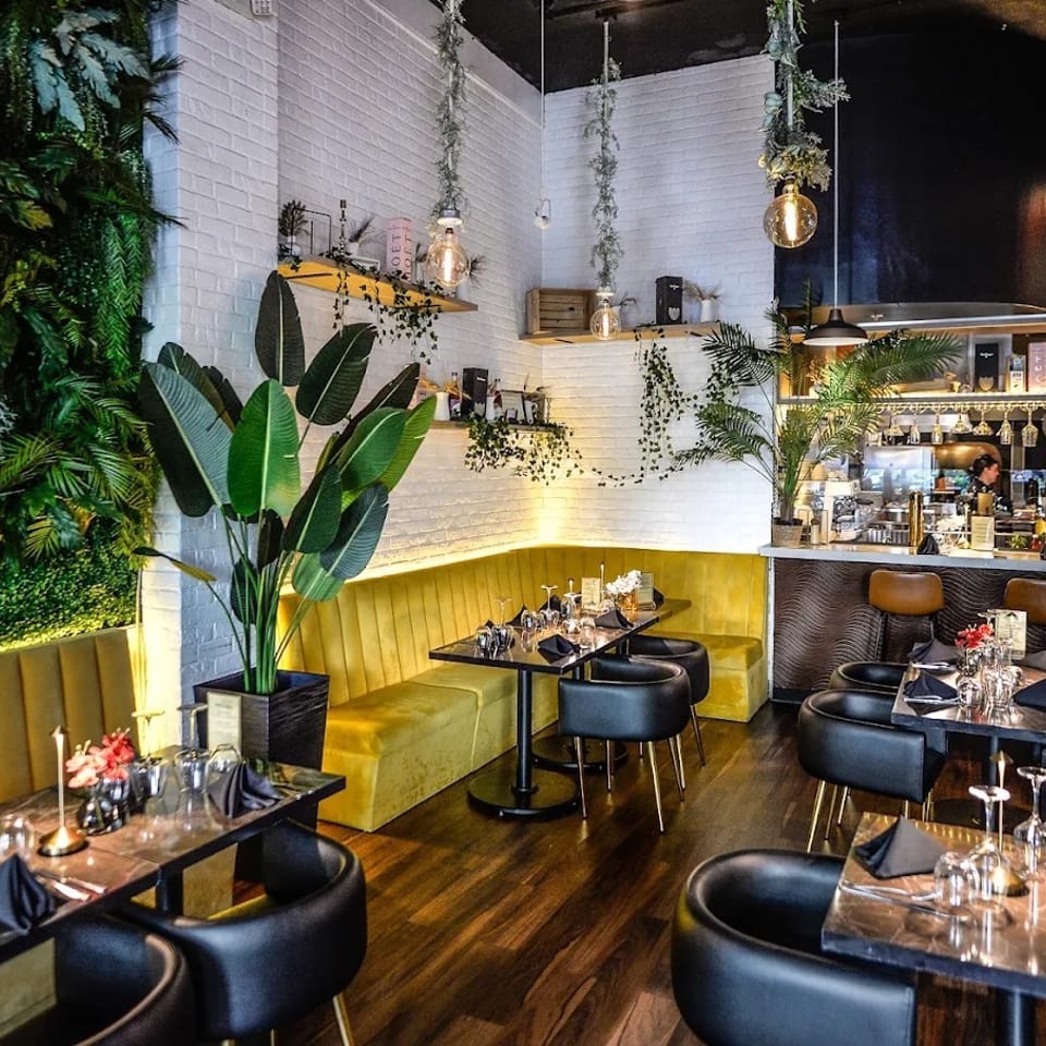 Elegant restaurant interior with black dining tables, mustard banquettes, lush greenery, warm pendant lights, and a polished bar.