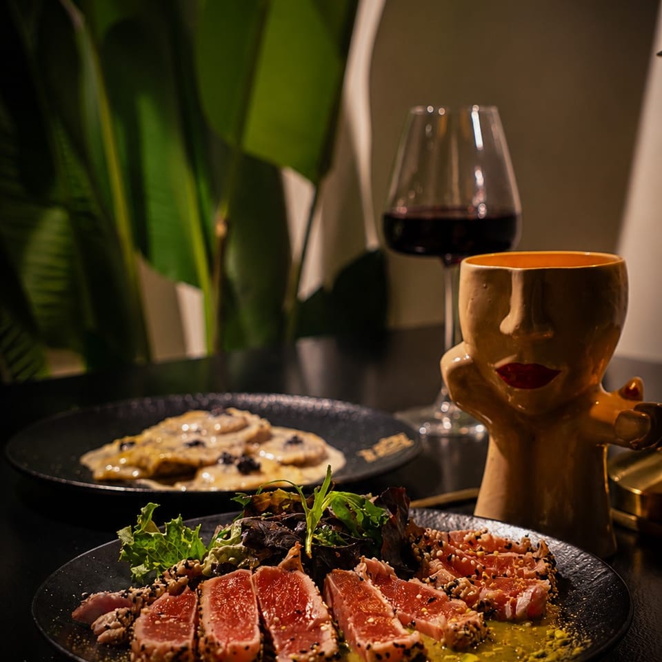 Seared tuna slices with salad and sauce on black plates, accompanied by red wine and a whimsical ceramic mug in a dim restaurant setting.
